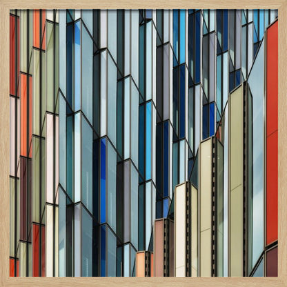 A close-up abstract photograph of a modern building's facade, featuring a complex pattern of colorful, angular glass panels in shades of blue, orange, red, and gray, all presented within a light wood frame. Wall Art