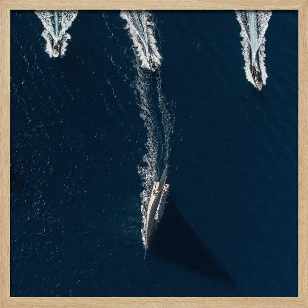 An aerial photograph from directly above shows a large sailboat and three smaller motorboats moving across a deep blue sea, leaving white wakes behind them. The sailboat is in the foreground, casting a long shadow, while the smaller boats are positioned further back. The entire image is enclosed in a light-colored wooden frame. Wall Art