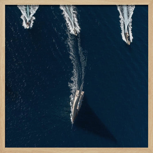 An aerial photograph from directly above shows a large sailboat and three smaller motorboats moving across a deep blue sea, leaving white wakes behind them. The sailboat is in the foreground, casting a long shadow, while the smaller boats are positioned further back. The entire image is enclosed in a light-colored wooden frame. Wall Art