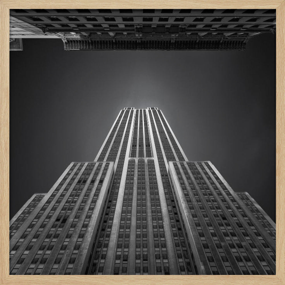 A dramatic, low-angle, black and white photograph of the Empire State Building, looking up from the street. The building's Art Deco lines stretch towards a dark grey sky, with another building visible at the very top of the frame, all enclosed in a light brown wood frame. Wall Art