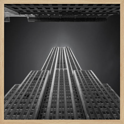 A dramatic, low-angle, black and white photograph of the Empire State Building, looking up from the street. The building's Art Deco lines stretch towards a dark grey sky, with another building visible at the very top of the frame, all enclosed in a light brown wood frame. Wall Art