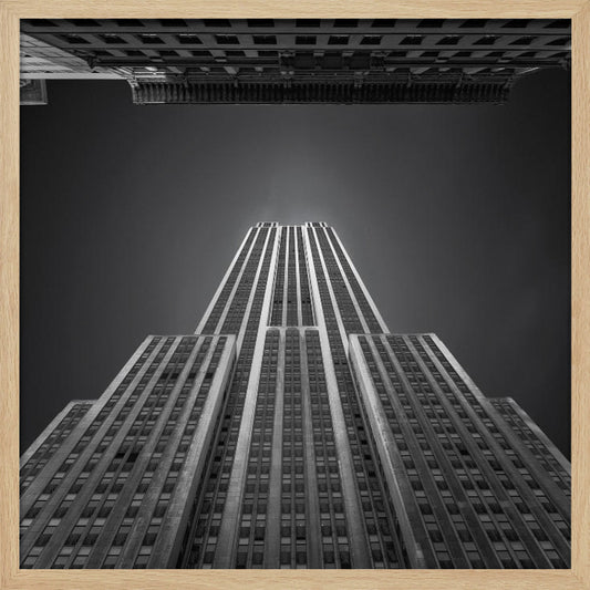 A dramatic, low-angle, black and white photograph of the Empire State Building, looking up from the street. The building's Art Deco lines stretch towards a dark grey sky, with another building visible at the very top of the frame, all enclosed in a light brown wood frame. Wall Art