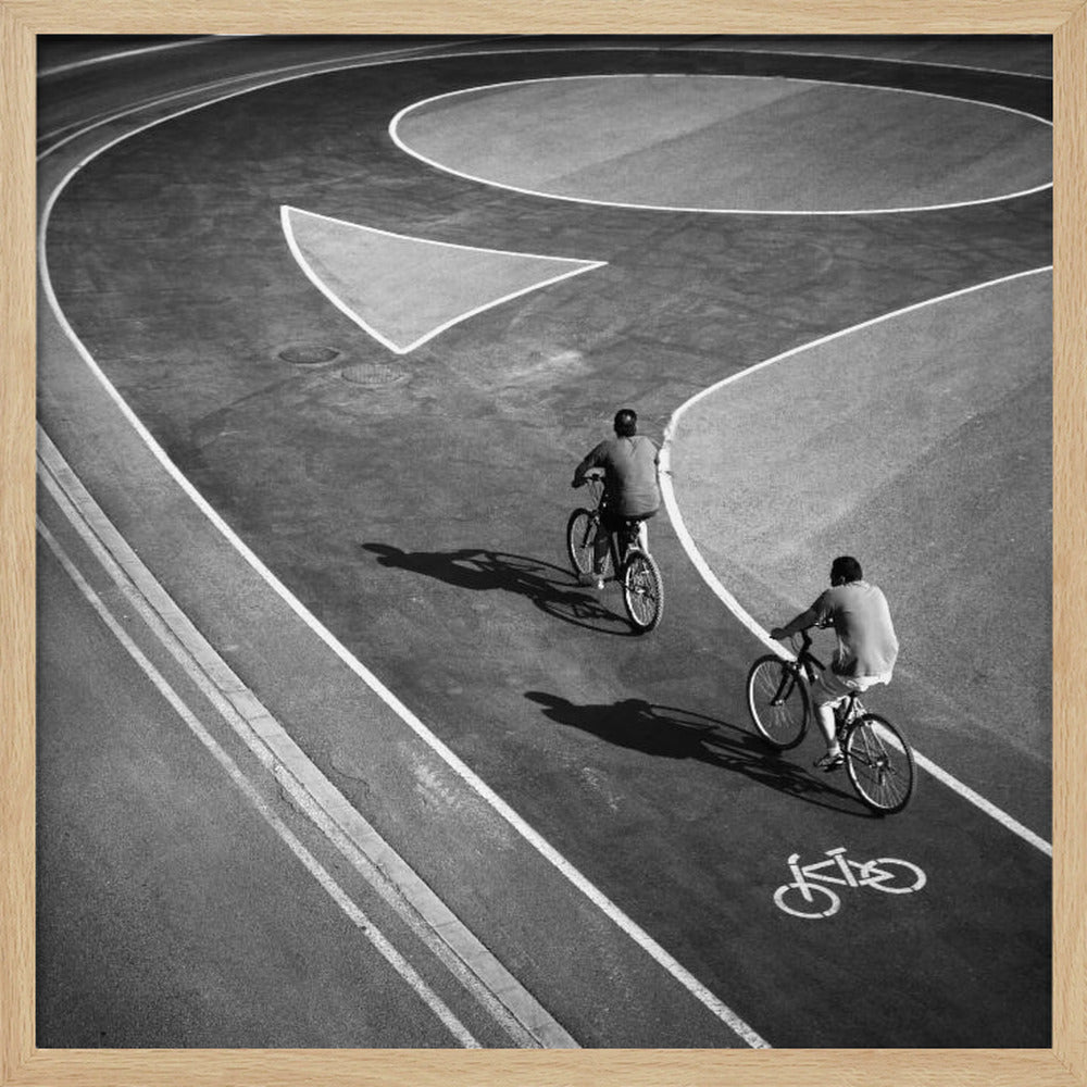 A framed, high-angle black and white photograph of two people cycling on a curving asphalt bike path. The path is marked with bold white lines, including a large arrow and a bicycle symbol, creating a strong graphic pattern. The cyclists cast long shadows on the pavement. Poster