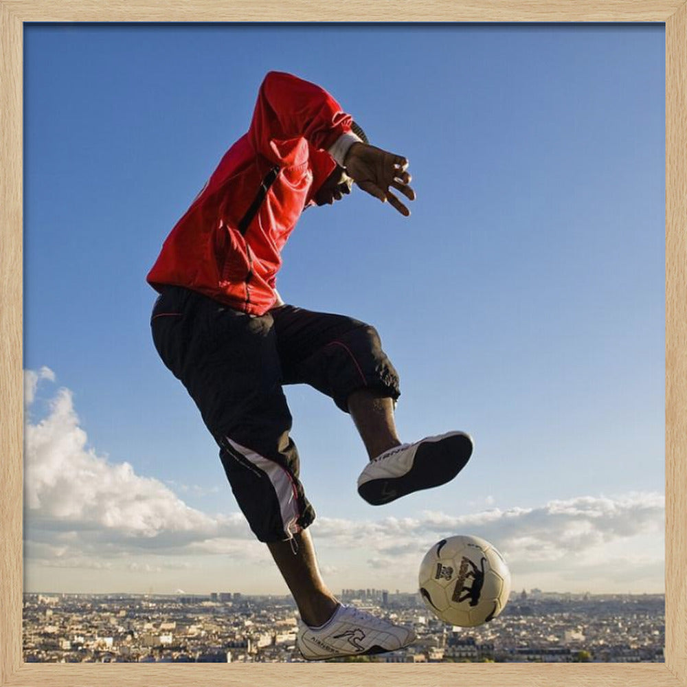 A dynamic low-angle shot of a person in a red jacket and black pants skillfully juggling a soccer ball, suspended in mid-air high above a sprawling city skyline under a clear blue sky, all enclosed within a light wood frame. Artwork