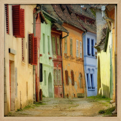 A narrow cobblestone street winds through a historic European town, flanked by colorful, weathered buildings in shades of cream, green, orange, and blue with red shutters, all under aged tile roofs and enclosed in a light wood frame. Artwork