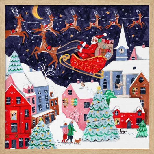 A festive illustration of Santa Claus flying in his sleigh pulled by reindeer over a snowy European-style village at night. The sky is a deep starry blue with a crescent moon, and the houses below are painted in vibrant reds, pinks, and blues, with warm light in the windows. People walk in the snow near decorated Christmas trees. Decor