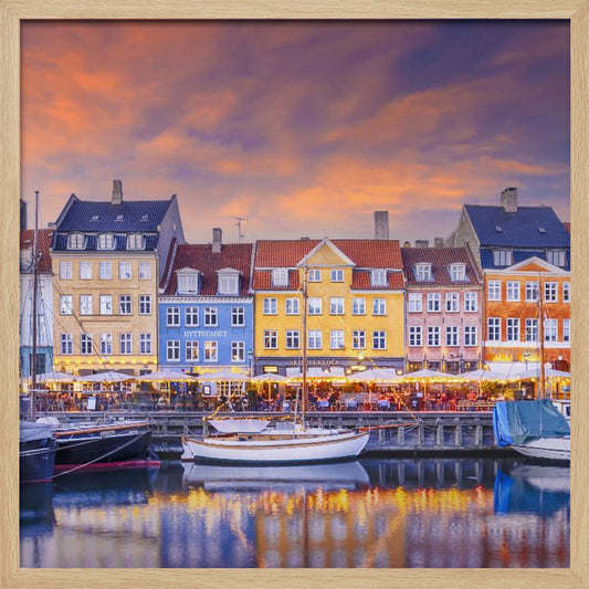 A framed photograph of the Nyhavn harbor in Copenhagen at dusk, featuring a row of brightly colored historic buildings along the canal. Sailboats are moored in the water, which reflects the warm glow of the building lights and the vibrant orange and purple sunset sky. Print