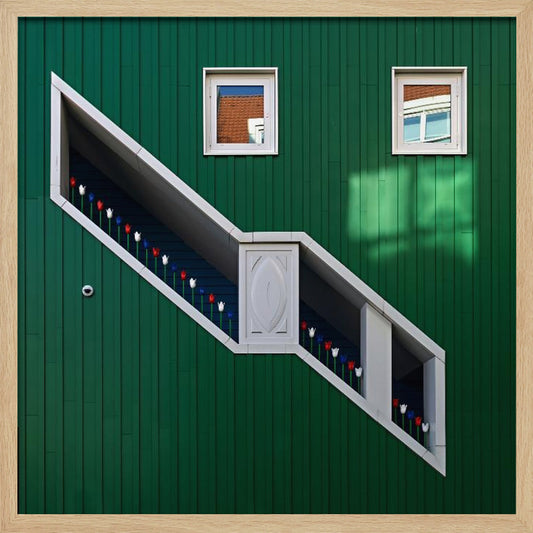 A framed photograph of a modern building with a dark green vertical-paneled facade, featuring a large, white-framed diagonal window that resembles a staircase, decorated with small red, white, and blue tulips. Two small square windows are positioned above. Decor