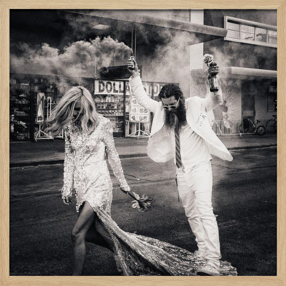 A black and white photograph in a light wood frame showing a stylish couple walking down a street. The woman wears a sparkling sequin gown, and the man, in a white suit with a long beard, triumphantly raises his arms, holding a smoke bomb and a bottle with a rose. Decor