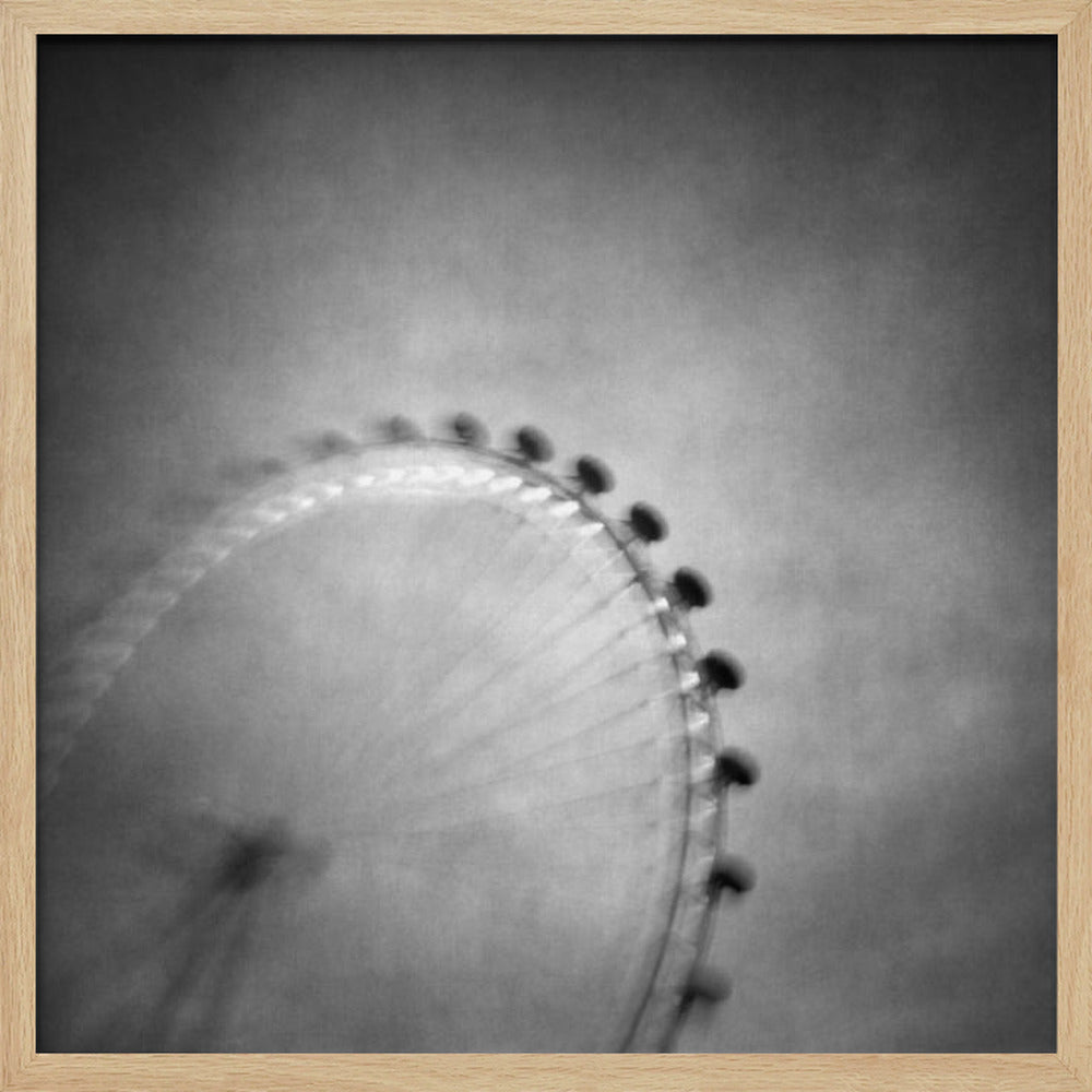 A moody, black and white long-exposure photograph of a large Ferris wheel, creating a soft, blurry effect of its rotation against a grainy gray sky. The artwork is presented in a simple, light wood frame. Poster