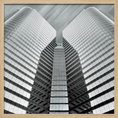 A low-angle, symmetrical black and white photograph of a modern glass skyscraper. The two towers of the building converge towards the center, creating a V-shape against a sky with motion-blurred clouds. The photo is enclosed in a light-colored wooden frame. Print