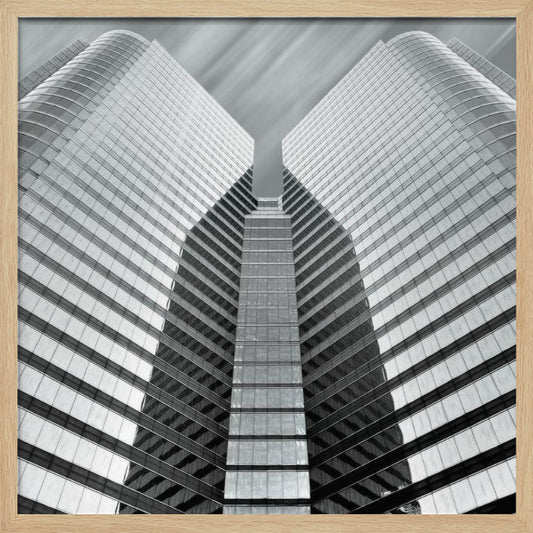 A low-angle, symmetrical black and white photograph of a modern glass skyscraper. The two towers of the building converge towards the center, creating a V-shape against a sky with motion-blurred clouds. The photo is enclosed in a light-colored wooden frame. Print