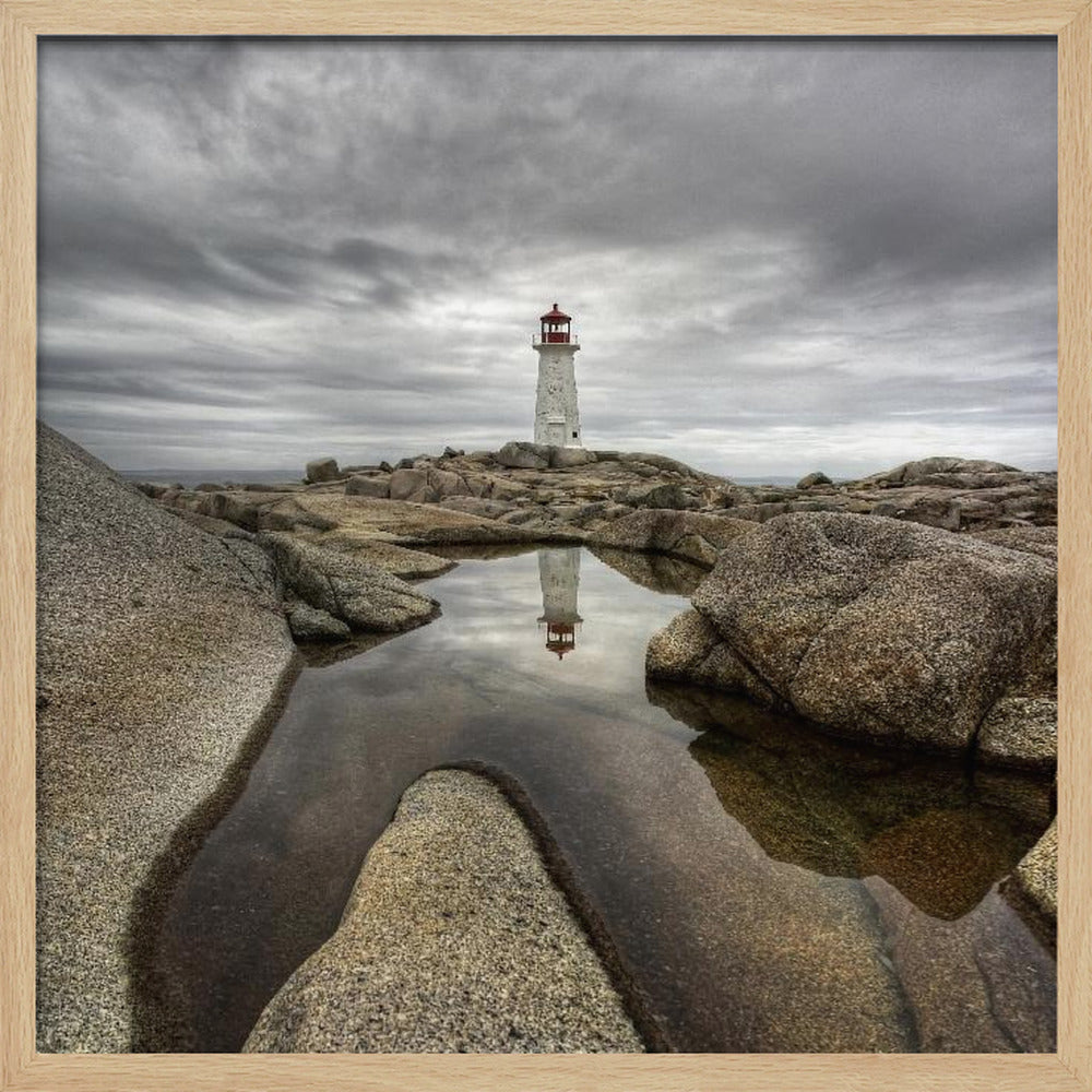 A framed photograph of a white lighthouse with a red top standing on a rocky shore under a dramatic, cloudy sky. In the foreground, a tide pool perfectly reflects the lighthouse. Artwork