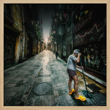 A wide-angle, atmospheric photograph of a man in bright yellow clogs mopping a wet, narrow cobblestone alley. The dark, gritty city street is lined with old buildings and closed storefronts, with another person walking in the distant light at the end of the alley. Wall Art