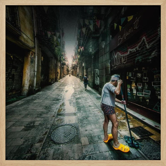 A wide-angle, atmospheric photograph of a man in bright yellow clogs mopping a wet, narrow cobblestone alley. The dark, gritty city street is lined with old buildings and closed storefronts, with another person walking in the distant light at the end of the alley. Wall Art
