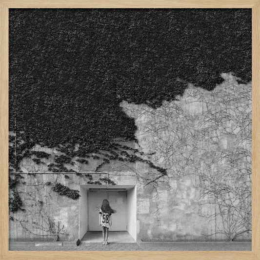A framed, square black and white photograph showing a stark contrast on an ivy-covered wall. The top half is dense with dark leaves, while the bottom half is a lighter, textured wall. A young girl with long hair, seen from behind, stands in a recessed white doorway in the center. Wall Art