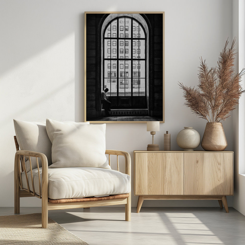 A black and white photograph of a solitary man sitting with his head bowed on the ledge of a massive arched window. The window's dark mullions create a grid over the bright view of another building outside, while the man remains in the shadows, creating a pensive and lonely mood. Poster