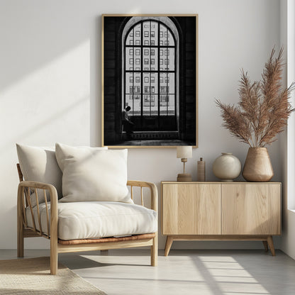A black and white photograph of a solitary man sitting with his head bowed on the ledge of a massive arched window. The window's dark mullions create a grid over the bright view of another building outside, while the man remains in the shadows, creating a pensive and lonely mood. Poster