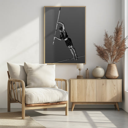 A dynamic black and white, low-angle shot of a female pole vaulter in mid-action. She is suspended in the air, gripping the pole as she prepares to clear the high bar, with her muscular form silhouetted against a plain sky. Print