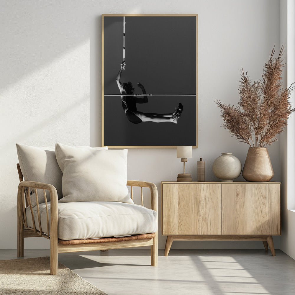 A dramatic black and white photograph of a male pole vaulter in mid-air, successfully clearing the horizontal bar. The athlete's body is parallel to the ground, with one arm still holding the vertical pole, against a solid gray sky. The image is enclosed in a simple black frame. Artwork