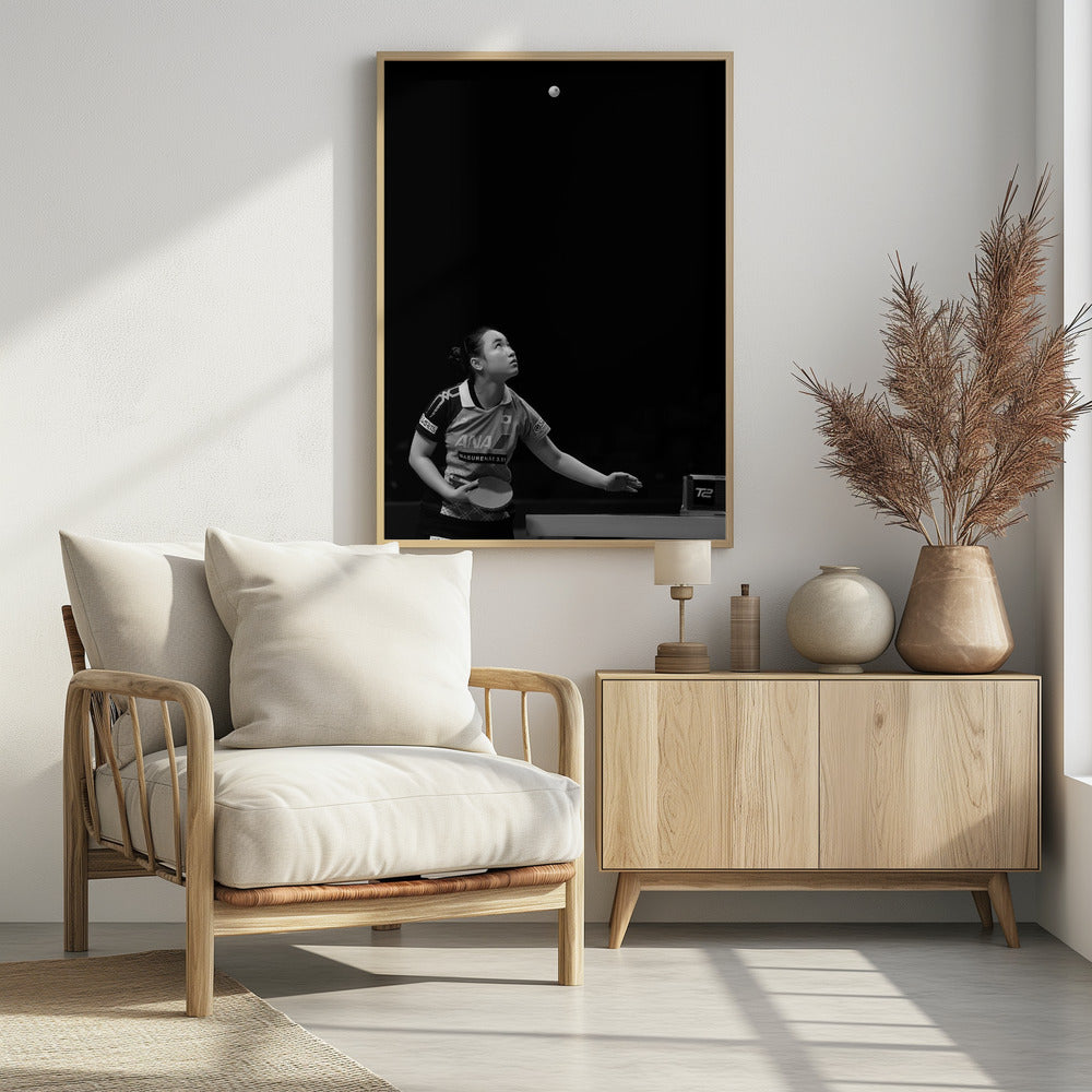 A dramatic black and white vertical photograph of a female table tennis player mid-serve. Positioned in the lower-left, she looks up with intense focus at the white ball suspended high in the air against a solid black background. Poster