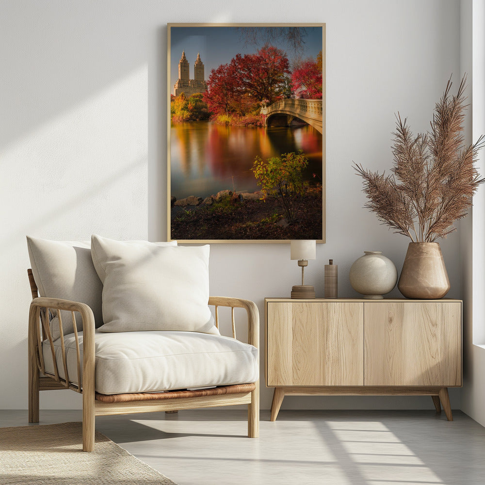 A picturesque autumn scene in Central Park featuring the Bow Bridge over a calm lake reflecting the vibrant red and yellow fall foliage, with the twin towers of The San Remo building visible in the background against a clear blue sky. Decor