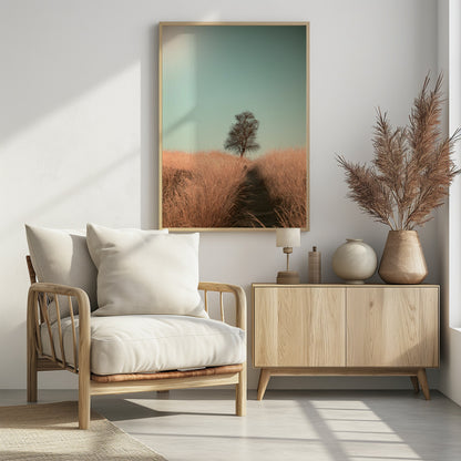 A minimalist photograph of a single bare tree standing alone on a gentle slope in a field of golden, dried grass under a clear, pale teal sky. A narrow path winds its way through the tall grass toward the tree. Print