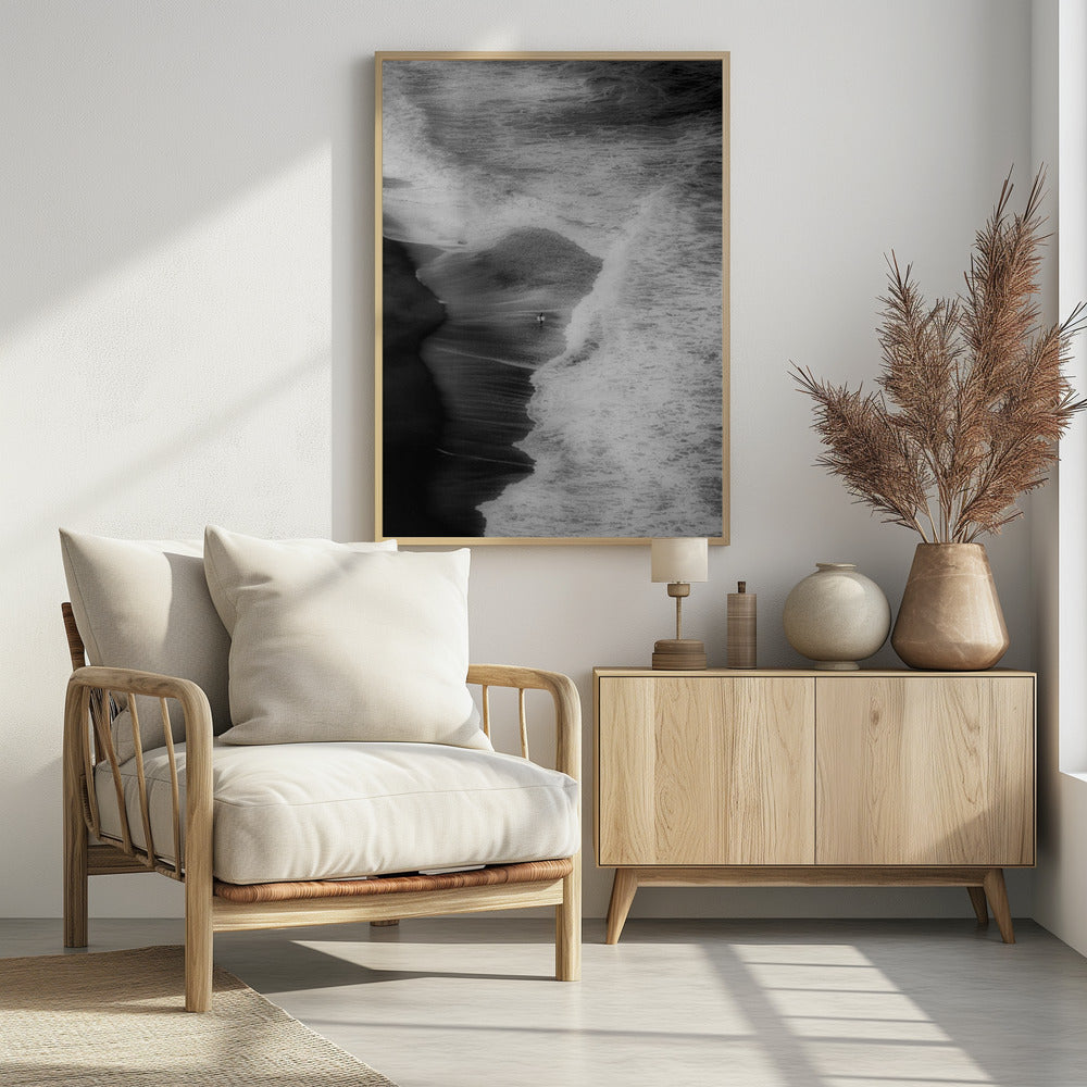 A high-angle, black and white photograph of a lone surfer carrying a surfboard at the water's edge on a dark sand beach. The vast, textured ocean with large, foamy waves crashing ashore dwarfs the solitary figure, creating a sense of scale and solitude. Decor