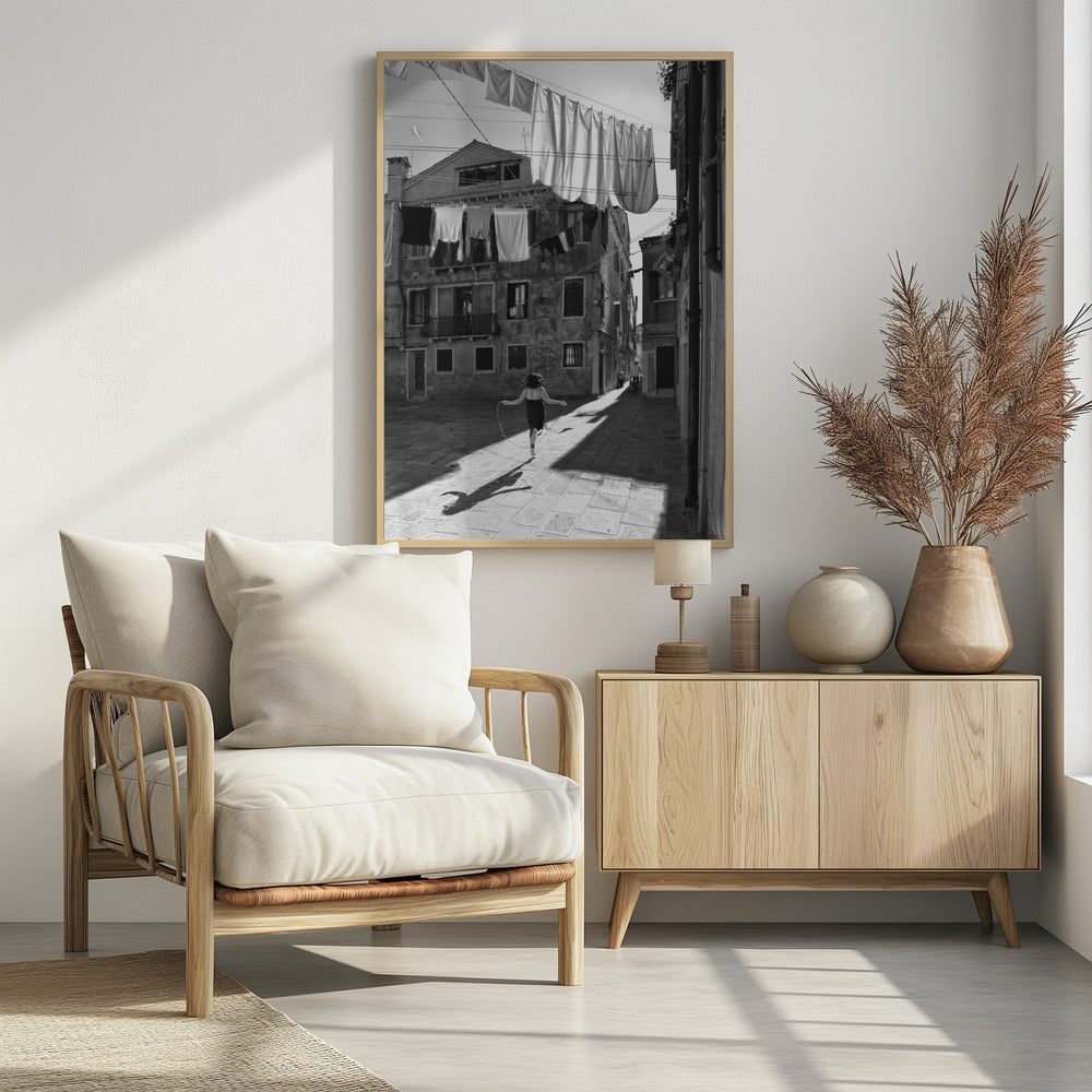A black and white photograph capturing a girl from behind as she jump-ropes down a sunlit, narrow cobblestone street in Venice. Her long shadow is cast before her, and above, laundry hangs on clotheslines strung between old, weathered European buildings. Wall Art