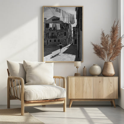 A black and white photograph capturing a girl from behind as she jump-ropes down a sunlit, narrow cobblestone street in Venice. Her long shadow is cast before her, and above, laundry hangs on clotheslines strung between old, weathered European buildings. Wall Art