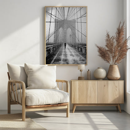 A vertical, black and white photograph of a solitary person walking along the Brooklyn Bridge during a heavy snowstorm. The iconic cables and towers of the bridge create a strong sense of perspective, drawing the eye towards the distant, hazy towers while snow falls all around. Artwork