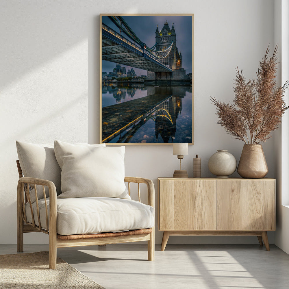 A dramatic, low-angle photograph of London's Tower Bridge at dusk. The iconic bridge is illuminated with warm yellow lights against a dark blue, cloudy sky. The calm River Thames in the foreground creates a stunning, clear reflection of the bridge and the distant city skyline. Print