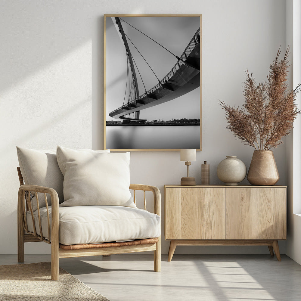 A dynamic low-angle, black and white photograph of a modern cable-stayed pedestrian bridge. The bridge curves elegantly over calm, reflective water, with its deck suspended by numerous cables attached to a tall, angled pylon, creating a striking graphic composition against a bright, clear sky. Decor