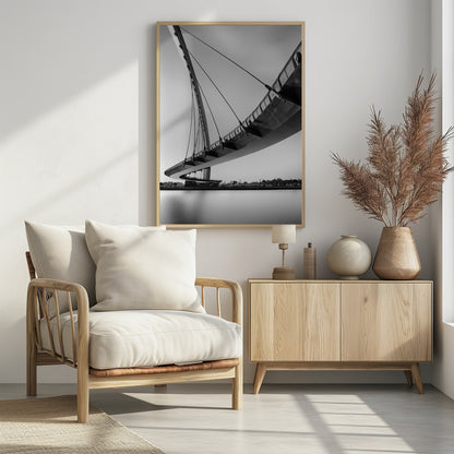A dynamic low-angle, black and white photograph of a modern cable-stayed pedestrian bridge. The bridge curves elegantly over calm, reflective water, with its deck suspended by numerous cables attached to a tall, angled pylon, creating a striking graphic composition against a bright, clear sky. Decor