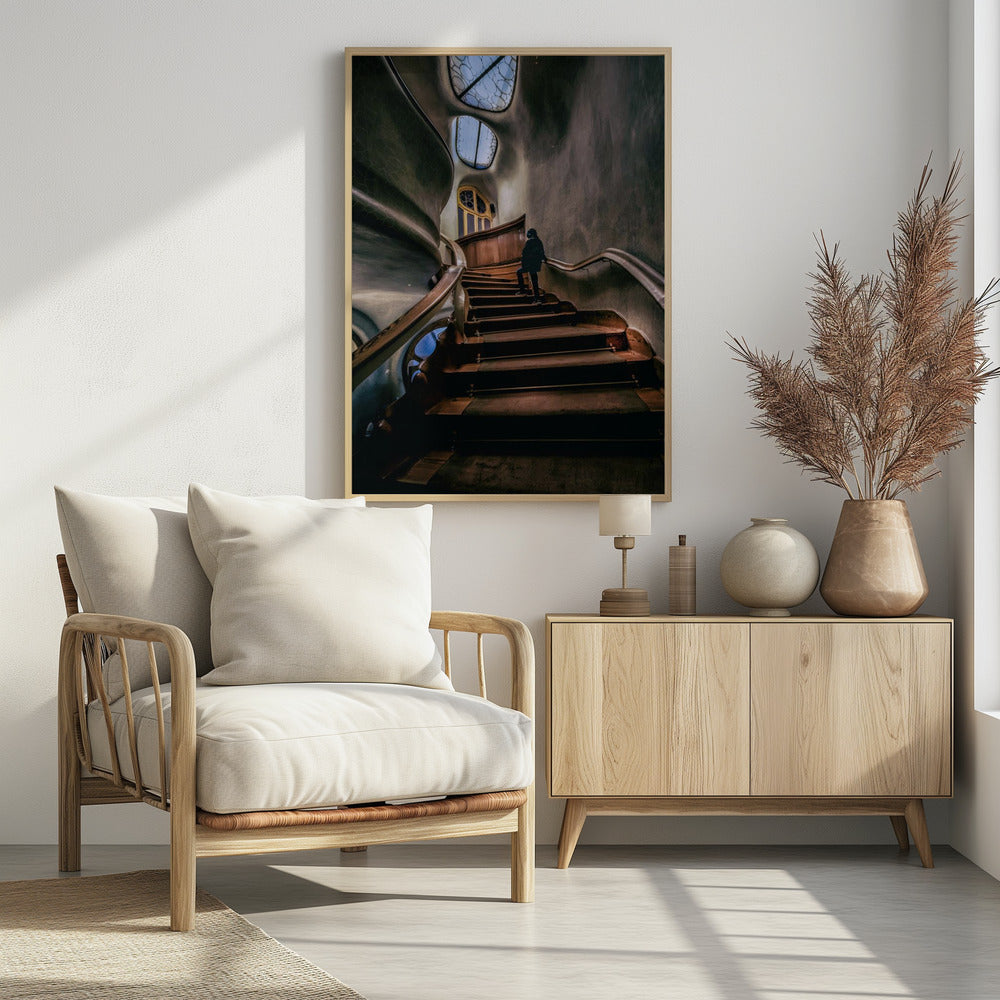 A low-angle view up a dark, winding wooden staircase inside a building with surreal, organic architecture. A person in a dark coat is walking up the stairs towards uniquely shaped windows that show a blue sky, contrasting with the dimly lit, textured gray walls. Print