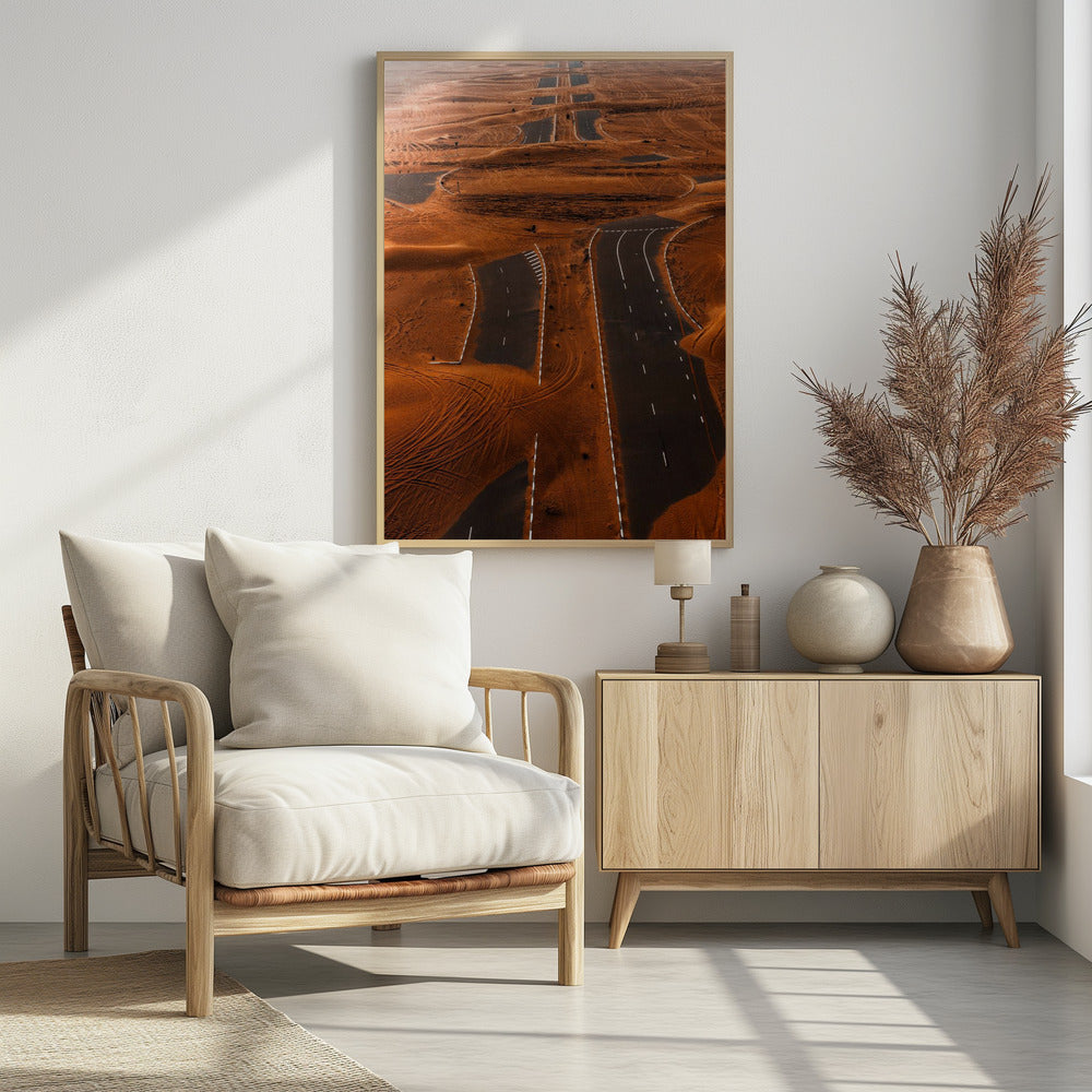 An aerial view of a multi-lane, black asphalt road being consumed by vast, rolling sand dunes of a deep orange color. The sand covers sections of the road, creating a surreal pattern of man-made lines and natural, windswept textures. Print