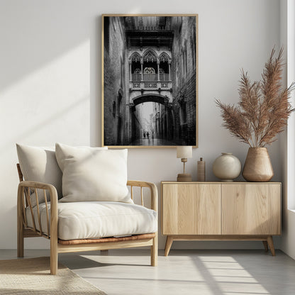 A moody, vertical black and white photograph of an ornate Gothic-style bridge connecting two stone buildings over a narrow alleyway. Below the arch of the bridge, the wet street stretches into the distance where two figures are walking away. Decor