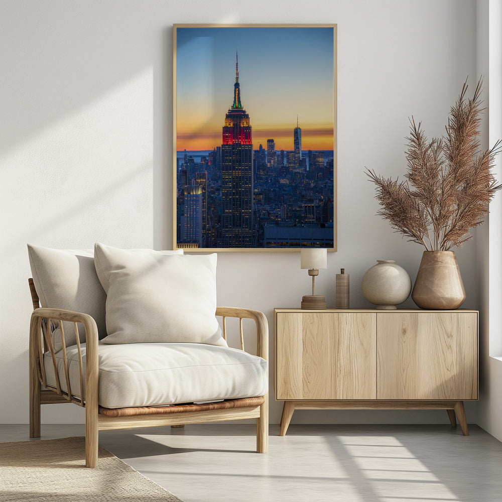 A vertical photograph of the New York City skyline at sunset, with the Empire State Building in the foreground. The building is illuminated with festive red and green lights on its upper floors, set against a sky that transitions from deep blue to a warm orange glow at the horizon. The surrounding cityscape, including the distant One World Trade Center, is bathed in the cool light of dusk, with scattered warm lights from building windows. Decor