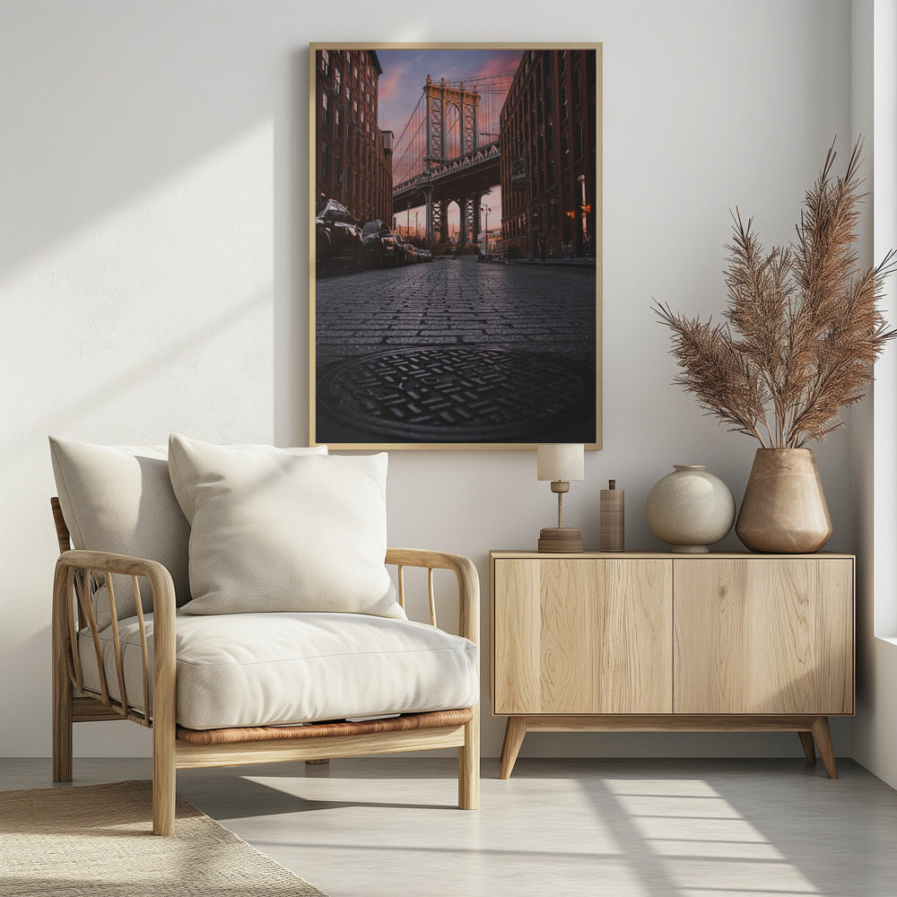 A low-angle photograph taken from a cobblestone street in Dumbo, Brooklyn, looking up towards the Manhattan Bridge at sunset. Red brick buildings frame the view on both sides of the street, with parked cars lining the left. A manhole cover is prominent in the foreground, and the sky is filled with warm pink and orange tones. Print