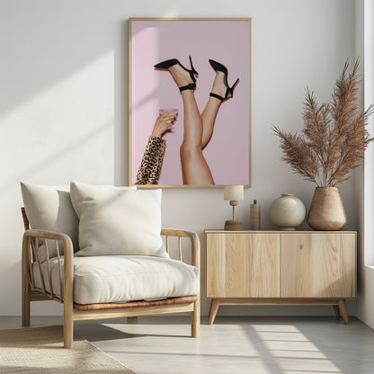 A woman's legs are kicked up in the air against a solid pink background. She is wearing black, pointed-toe, high-heeled shoes with ankle straps. A hand in a leopard print sleeve holds a pink, textured cocktail glass to the side. Artwork