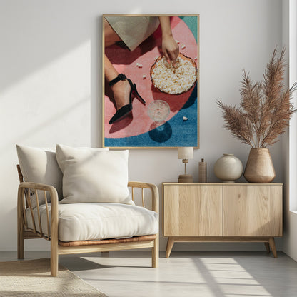 A high-angle photograph of a woman sitting on a vibrant pink and blue rug. She is wearing a chic black stiletto high heel and reaching into a glass bowl filled with popcorn. A crystal glass and scattered popcorn kernels are also on the rug. Artwork