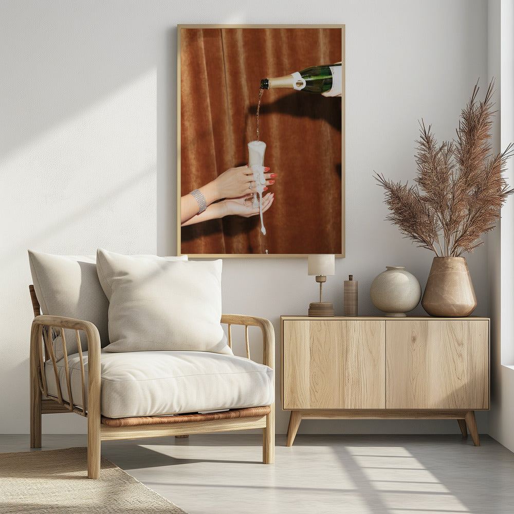 Champagne is poured from a bottle into a glass, overflowing with white foam that drips down onto two manicured hands with red nail polish holding it against a brown velvet curtain background. Poster