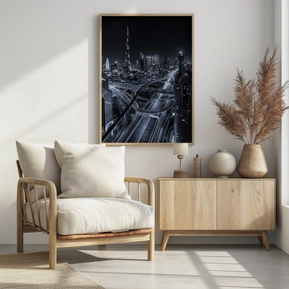 A cool-toned black and white photograph of the Dubai skyline at night. The iconic Burj Khalifa stands tall in the background, while the foreground is dominated by a massive, complex highway interchange with light trails from moving cars, creating a sense of motion and modernity. Poster