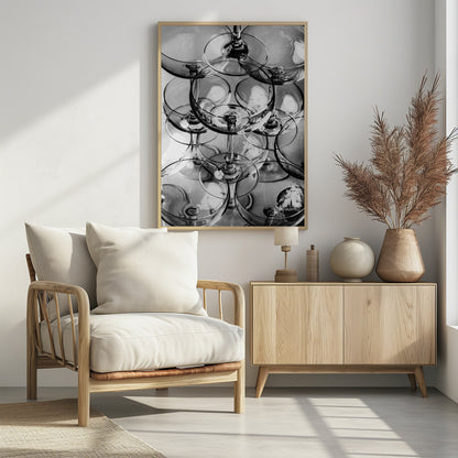 A black and white, overhead photograph of a tightly packed stack of empty champagne coupe glasses. The overlapping rims, stems, and bowls create a complex abstract pattern of circles and reflections, all enclosed within a simple black frame. Poster