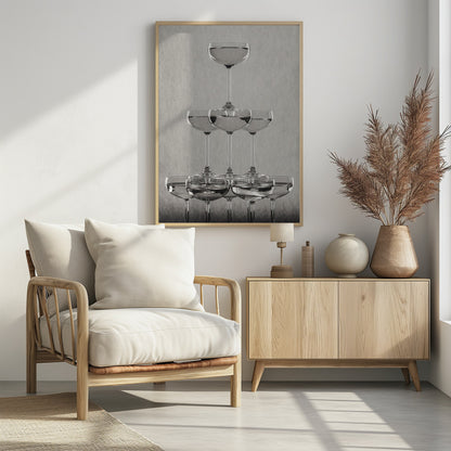 A classic black and white photograph of a champagne tower, with coupe glasses stacked in a pyramid. The glasses are filled with bubbly liquid, and the composition is set against a textured light gray background and enclosed in a black frame. Poster