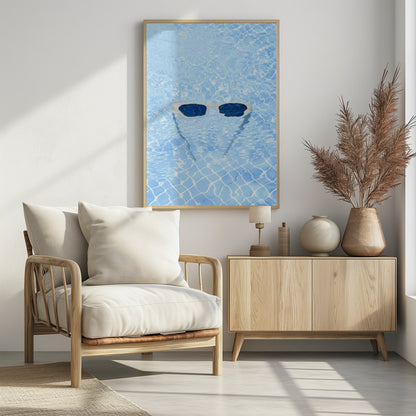 A pair of white-framed sunglasses with dark lenses floats on the surface of a bright blue swimming pool. The rippling water distorts the view of the tiled pool floor, and the sunglasses cast a faint shadow underneath. Print