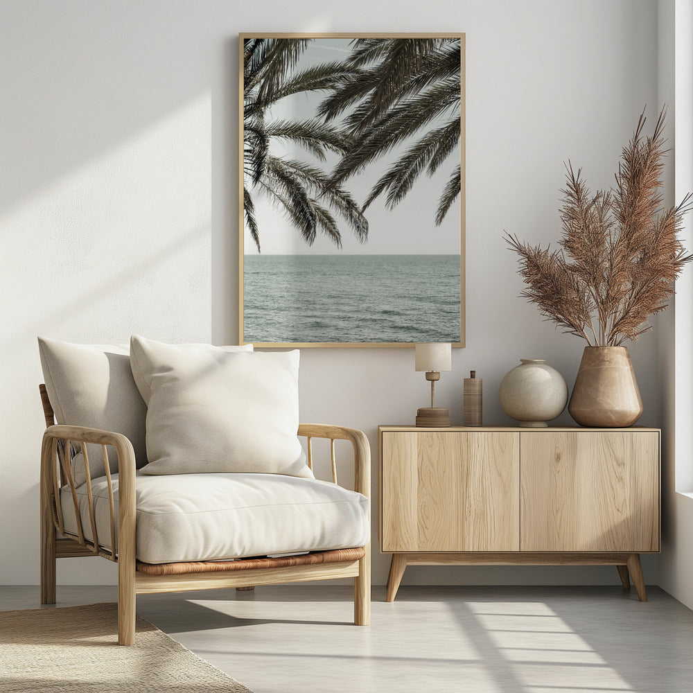 A close-up view of dark green palm tree fronds hanging over a calm, muted sage green ocean under a pale, overcast sky. The photograph has a serene and tranquil mood and is displayed in a black frame. Wall Art