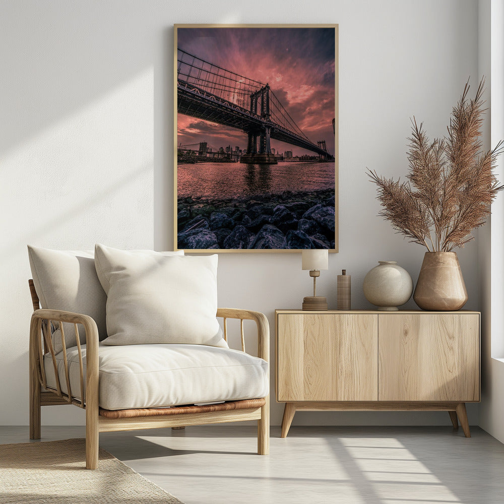 A dramatic low-angle view of the Manhattan Bridge over the East River at sunset. The sky is filled with pink and red clouds, which are reflected in the water below. The dark silhouette of the bridge contrasts with the vibrant sky, and the New York City skyline is visible in the background. In the foreground, dark, wet rocks line the riverbank. Decor