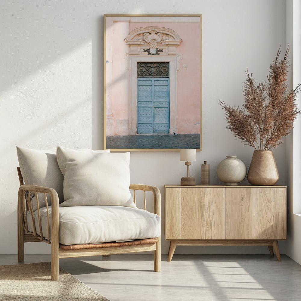 A light blue, studded double door set into a pale pink wall. The doorway is framed in white stone with an ornate pediment and a decorative wrought-iron transom window above the door. A single pigeon stands on the cobblestone pavement in front. Print