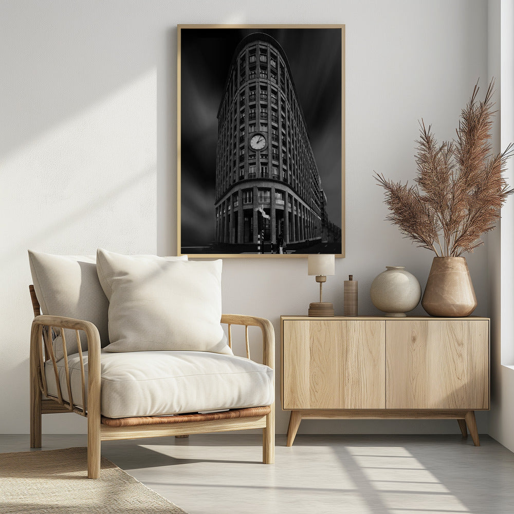 A dramatic low-angle, black and white photograph of a large, curved corner building with a prominent clock on its facade. The sky is dark with a motion blur effect, making the building appear grand and imposing. Artwork